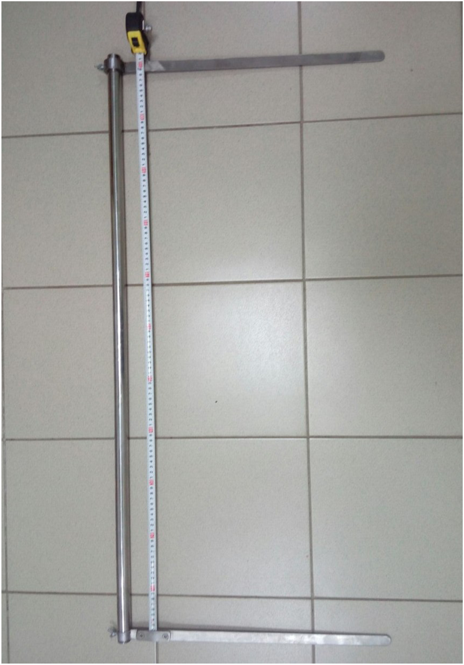 Measuring device consisting of a tape measure attached to a metal frame, with horizontal and vertical arms positioned on a tiled floor.