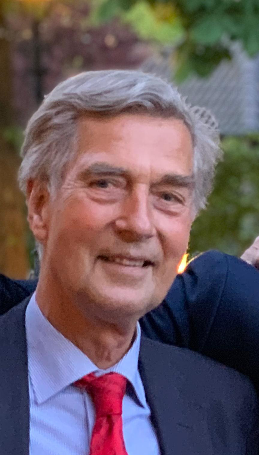An older man with gray hair is smiling at the camera. He is wearing a dark suit, a light blue shirt, and a bright red tie. The background shows blurred greenery.